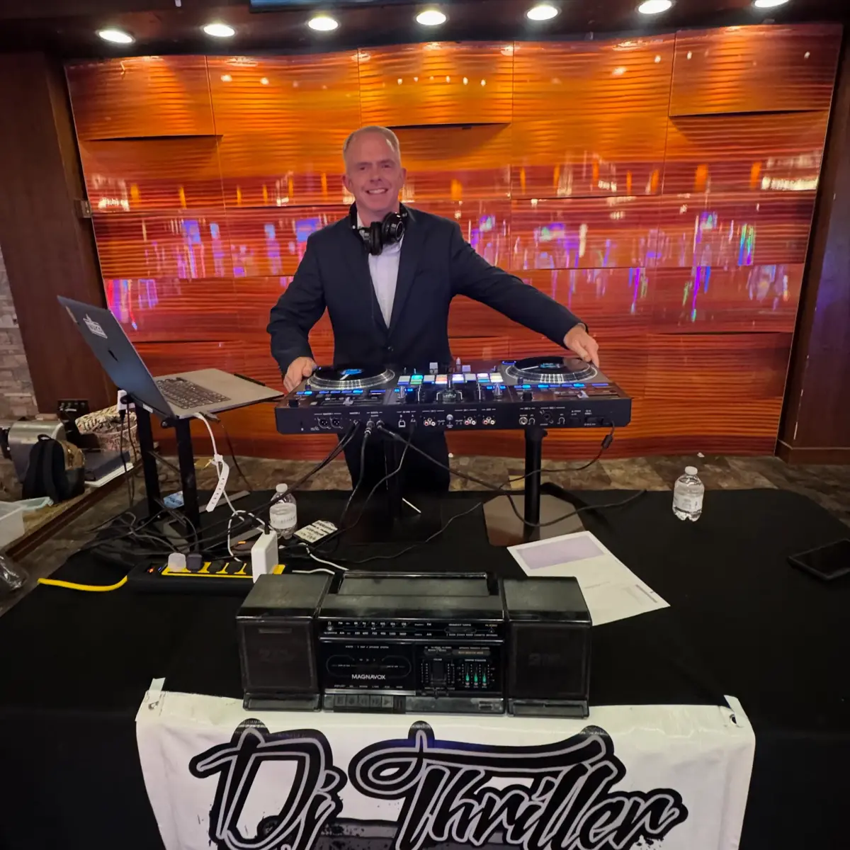 DJ Thriller in navy blazer behind professional DJ controller setup at indoor venue with warm copper lighting